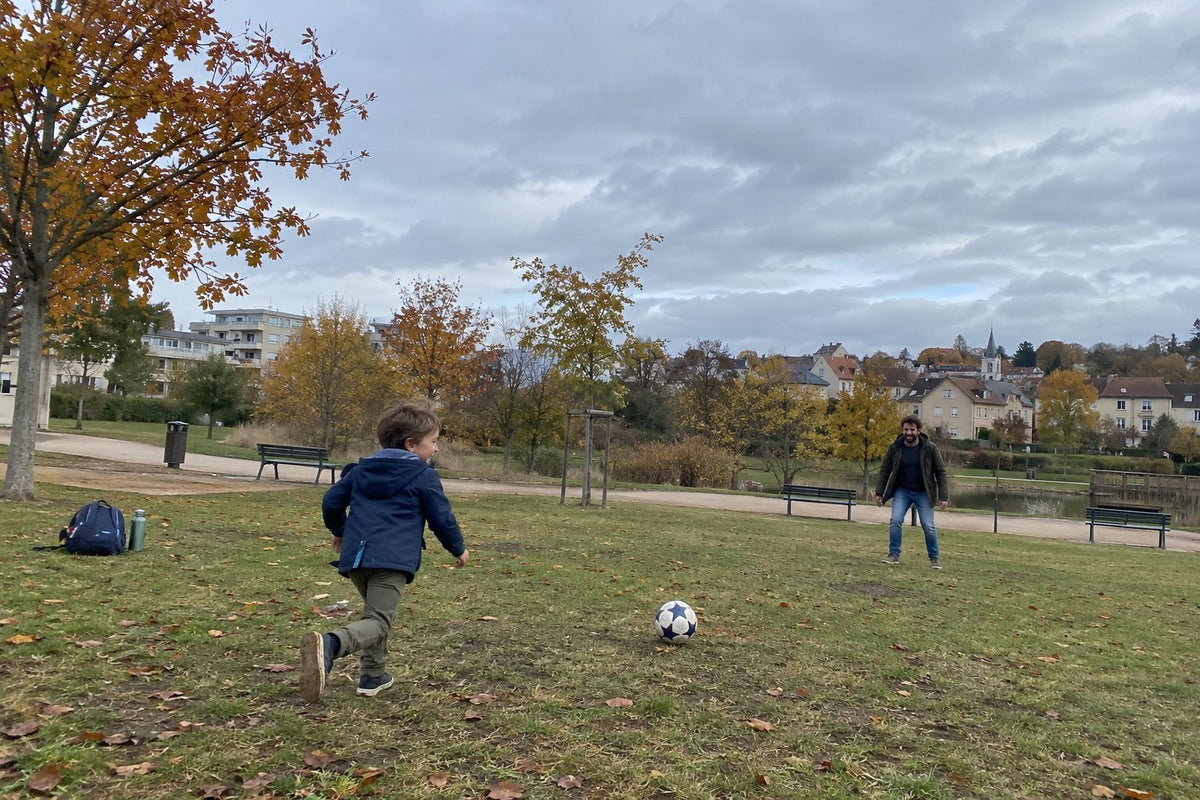 Un enfant court vers le ballon dans un parc en automne, son père joue avec lui