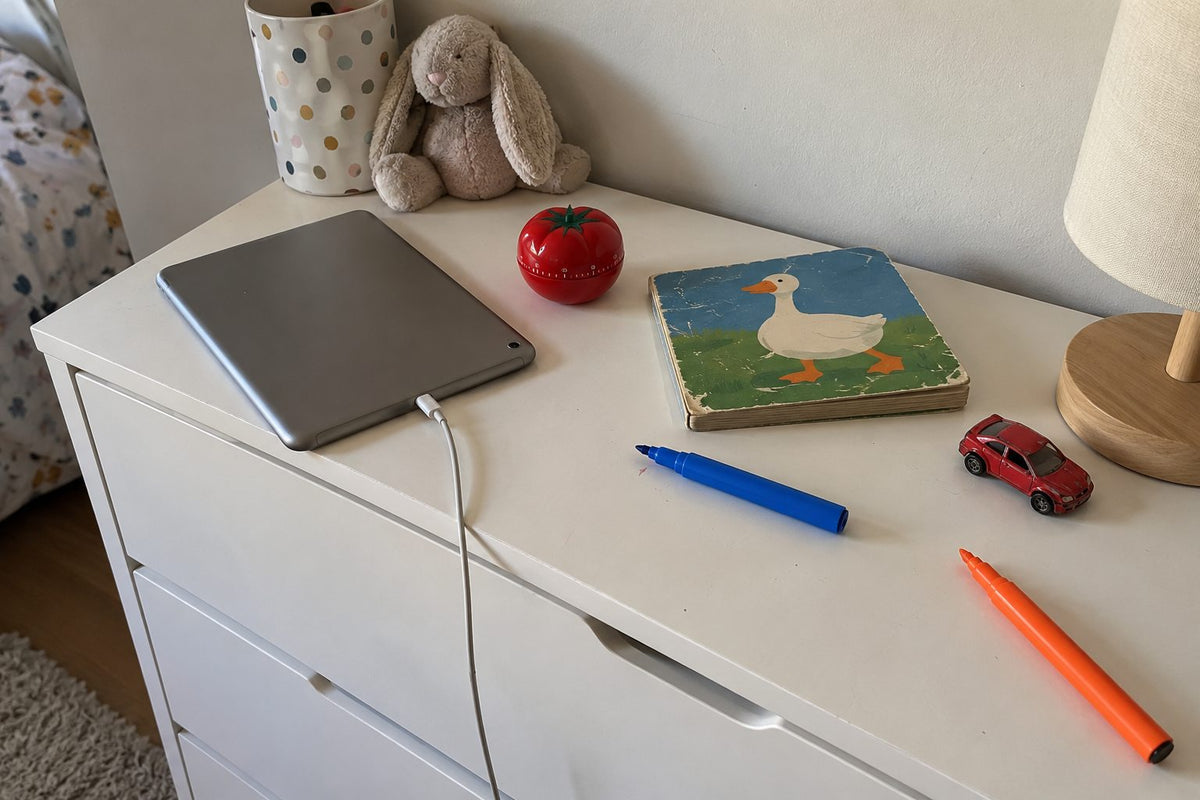 Une tablette retournée en charge, un minuteur et un livre d’enfant sur une commode de chambre